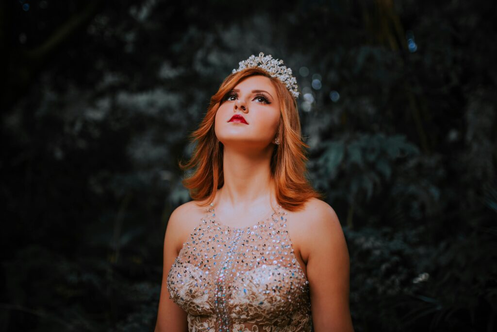 Enchanting portrait of a woman with red hair wearing a tiara and a glittering dress, gazing upward in a forest.